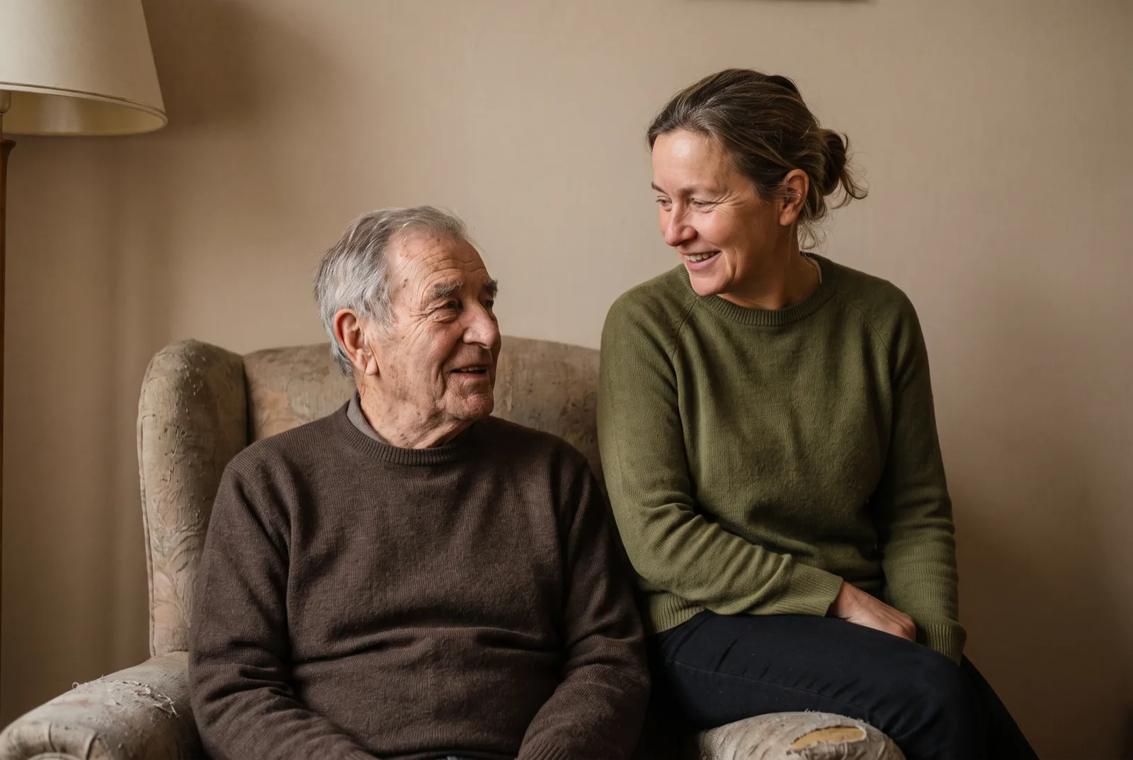 An elderly man in an armchair talking with his adult daughter seated beside him